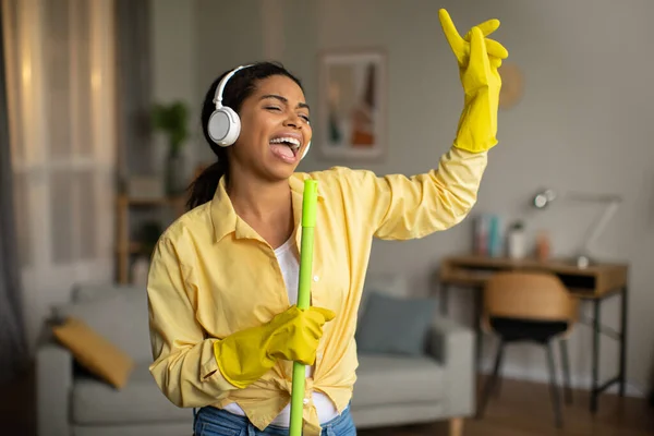 Cheerful Black Female Cleaning House Wearing Rubber Gloves And Singing Having Fun Holding Mop Like Microphone Posing Standing In Modern Living Room. Lady Enjoying Cleaning Home On Weekend