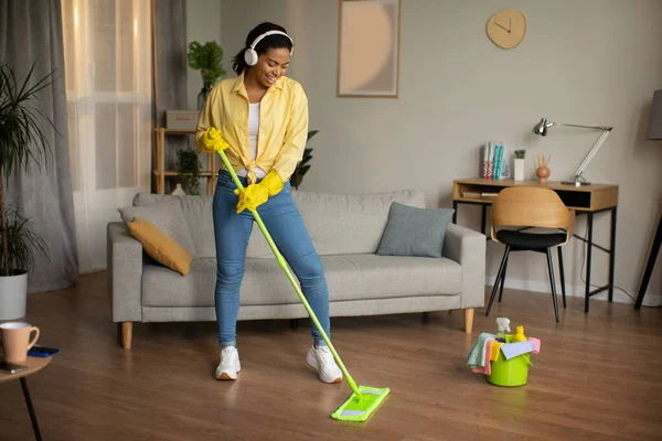 Cheerful Black Female Mopping Floor And Listening To Music In Wireless Headphones Enjoying House Chores Standing In Modern Living Room At Home. Lady Cleaning House. Housework Concept