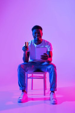 İnternetten alışveriş. Dizüstü bilgisayarı ve kredi kartı olan neşeli siyah bir adamın portresi. Sandalyede oturuyor, uzun boylu. Neşeli Afrikalı Amerikalı adam internetten ödeme yapıyor, nakit para alıyor.