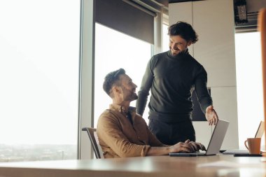 Gülümseyen şirket çalışanları iş birliği yapıyor, ortak iş projesinde çalışıyor, iş yerinde dizüstü bilgisayar kullanıyor, kopyalama alanı kullanıyor. Modern ofiste arkadaşça sohbet eden mutlu erkek arkadaşlar.