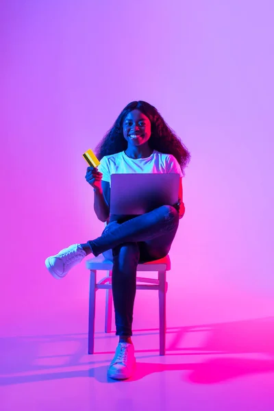 オンライン決済 クレジットカードとノートパソコンを持つ幸せなアフリカ系アメリカ人女性がネオンで椅子に座っています インターネットでリモートショッピングやお支払いを楽しむ楽しい若い黒人女性 — ストック写真