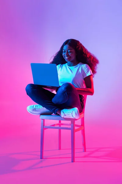 Happy young black woman using laptop pc for online work or studies, sitting on chair in neon light, full length. Cheery African American lady having remote video conference, chatting on web