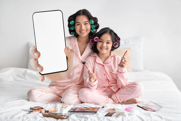 Beauty App. Asian mother and little daughter showing blank smartphone at camera, mom and child wearing silk pajamas holding mobile phone and doing makeup on bed, having fun at home, collage, mockup