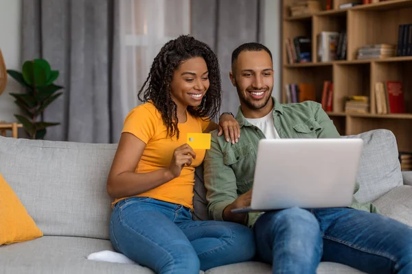 Online wallet and banking concept. Excited african american spouses using laptop and making purchases via internet store, paying with debit credit card, sitting on sofa at home