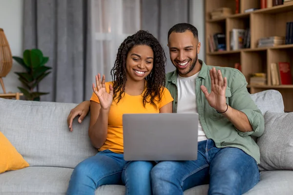Modern remote communication. Happy black couple making digital video call with friends or family using laptop, waving to webcam, sitting on couch at home