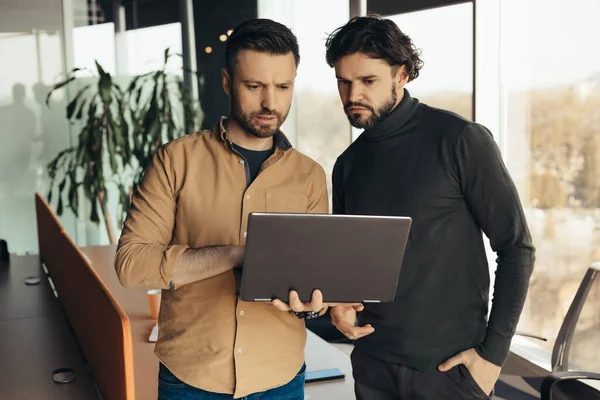 İki odaklanmış erkek çalışan iş stratejileri geliştiriyor, birbirleriyle işbirliği yapıyorlar, modern ofiste dizüstü bilgisayarlarını kullanıyorlar. Ciddi iş arkadaşları taşınabilir bilgisayarın yanında başlangıç fikirleri için beyin fırtınası yapıyorlar