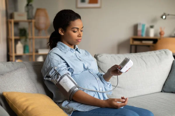怀孕的黑人女性在家中坐在沙发上测量低张力问题的血压 怀孕期间的健康问题概念 侧视拍摄 选择性聚焦 — 图库照片