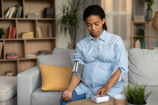 Pregnant African American Lady Measuring Arterial Blood Pressure Having Hypertension Problem Expecting Baby Sitting On Couch At Home. High And Low Tension Symptom Concept