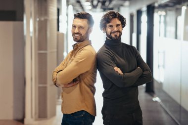 Kollarını kavuşturup şirket salonunda gülümseyen yakışıklı girişimcilerin portresi. Çağdaş ofiste sırt sırta vermiş güler yüzlü iş adamları. İş ortaklığı kavramı