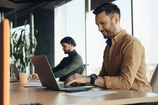 Profesyonel meslek, iş birliği konsepti. Dizüstü bilgisayar kullanan, e-postaları kontrol eden, klavyede yazan, açık uzay ofisinde çalışan mutlu bir erkek şirket çalışanı.