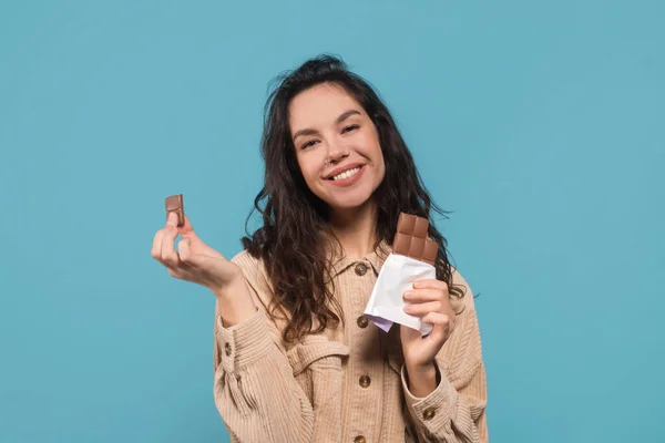 青の背景 スタジオに隔離されたカジュアルな食事チョコレートバーで幸せな千年の白人女性のブルネットの学生を満足させました 甘い歯とお菓子 広告や素晴らしいオファー 顔の表情 — ストック写真