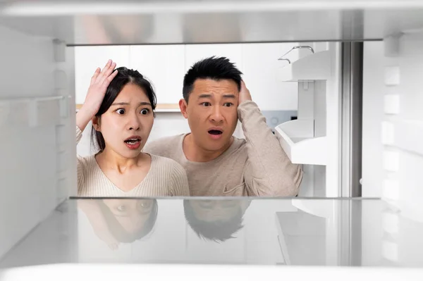 Confused chinese couple looking inside open empty fridge in kitchen at home, touching their heads. Poor asian family have nothing to eat, copy space. Financial hangover while crisis, poverty concept