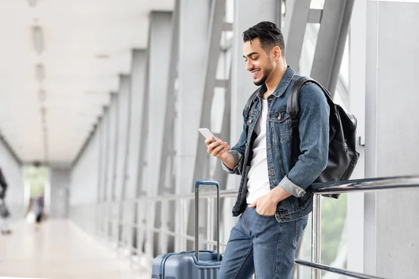 空港で待っているスマートフォンや荷物を持つ笑顔アラブ男性旅行者の肖像 端末に立っている間携帯電話でインターネットを閲覧幸せな若い中東の男 スペースをコピー — ストック写真