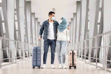 Pandemik Trip. Koruyucu tıbbi maskeler takan genç Müslüman çift havaalanında bavullarıyla, romantik İslami eşleri kucaklıyor ve birbirlerine bakıyor, birlikte yurtdışına seyahat ediyorlar.
