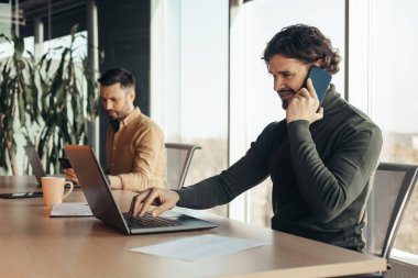 Genç ve neşeli iş adamı dizüstü bilgisayar kullanıyor, cep telefonuyla konuşuyor, şirket ofisindeki erkek arkadaşla çalışıyor, boş yer var. İş yerinde telefonla iletişim kuran yakışıklı milenyum çalışanı.