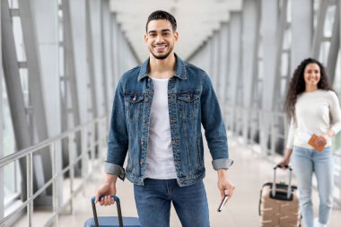 Hava Yolları. Havaalanında bavullu gülümseyen Arap adam, Mutlu Orta Doğu erkek yolcusu Terminal 'de çantasıyla duruyor ve biletlerle pasaportunu tutuyor,