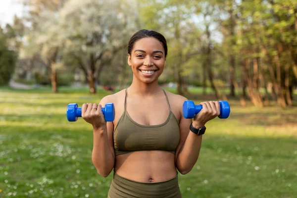 Cheery young black woman doing outdoor strength training, exercising with dumbbells, working out muscles at city park. Fit African American lady taking care of her body, leading active lifestyle