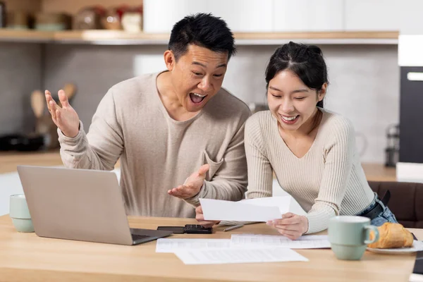 Happy asian couple paying bills online from home, sitting together at kitchen table, holding letters and papers, using computer and smiling, enjoying easy Internet banking, got good news, copy space