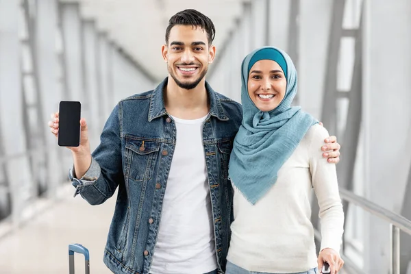 Travel App. Happy Muslim Couple Standing At Airport And Showing Blank Smartphone With Black Screen, Smiling Middle Eastern Spouses Advertising New Mobile Application For Flight Bookings, Mockup