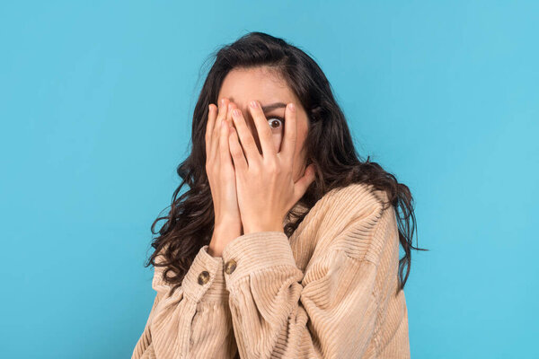 Scared millennial caucasian woman brunette student in casual covers her face with hands, isolated on blue background, studio, empty space, close up. Human emotions, facial expression, fear and afraid
