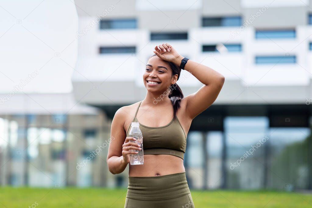 Tired young African American woman in sportswear taking break during jogging, wiping forehead, drinking water at city park. Millennial black lady keeping hydrated after outdoor workout