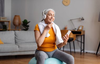 Omzunda havluyla ve kablosuz kulaklığıyla mutlu bir son sınıf kadını Fitball 'da oturuyor, cep telefonuyla müzik dinliyor, antrenman sırasında dinleniyor, boş bir alanda.