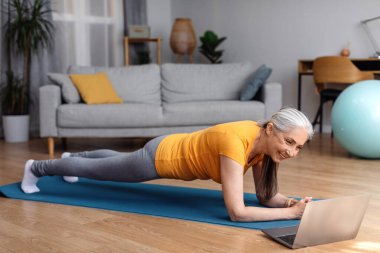 Aktif yaşlı kadın evde egzersiz yapıyor, dizüstü bilgisayarda spor videoları izliyor ve planking yapıyor, fitness dersi alıyor, kopya çekiyor. Sağlıklı yaşam tarzı kavramı