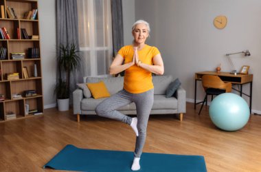 Emeklilik üzerine yoga pratiği. Sakin yaşlı bayan ağaç duruşu yapıyor, dengesini koruyor, evde yoga minderiyle egzersiz yapıyor. Evde antrenman yapan mutlu yaşlı kadın, kopyalama alanı