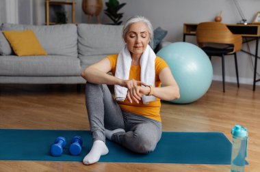 Kıdemli sporcu kadın, spor yaptıktan sonra spor takip cihazı kullanıyor. Yanmış kalorileri kontrol ediyor. Oturma odasının iç tarafındaki yoga minderinde oturuyor.