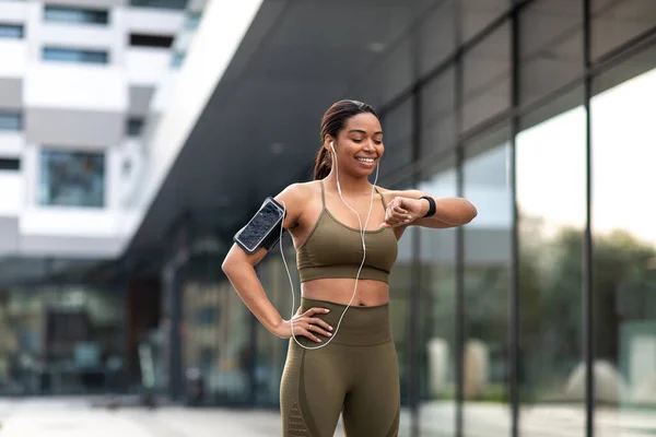 Young black woman looking at smartwatch, tracking fitness activity, listening to music in earphones during outdoor training. Fit African American female checking heart rate while jogging