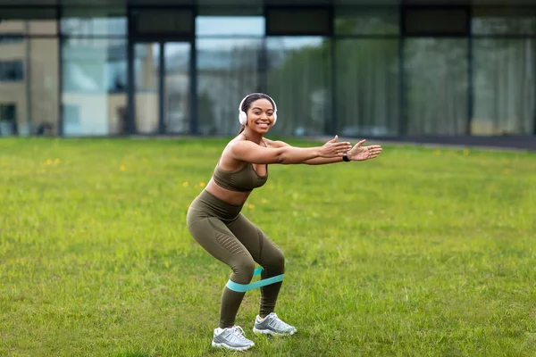 Sportif Afrikalı genç bir kadın elastik bantla çömelme hareketi yapıyor, kulaklıkla müzik dinliyor açık hava antrenmanı sırasında, boş alan. Şehir parkında bacak kaslarını çalıştıran siyahi bir kadın.