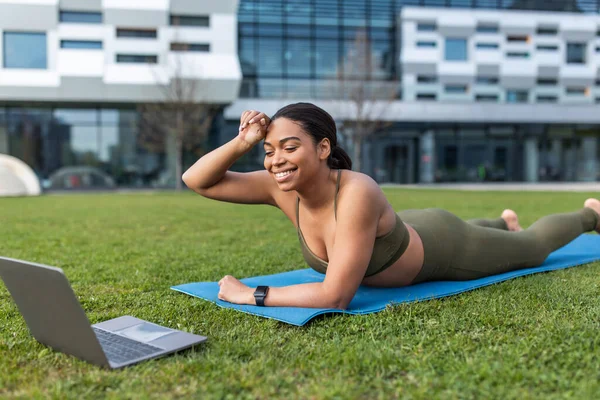 Tired young black woman lying on mat near laptop, taking break, wiping forehead during online training at urban park, free space. Millennial lady exercising outdoors, following video tutorial
