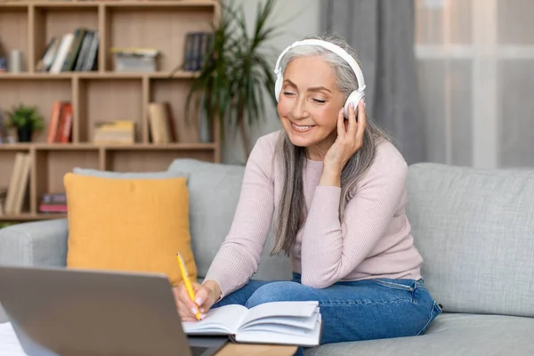 快乐漂亮的退休的欧洲老灰发女士在无线耳机与笔记本电脑在客厅内部工作 在家学习 在Covid 19大流行期间喜欢的音乐和网络研讨会 自由自在的空间 — 图库照片