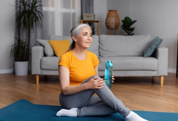 穿着运动服 身强体壮的老年女士 坐在健身垫上 喝着一瓶水 面带微笑 在家里锻炼时休息 自由自在 — 图库照片
