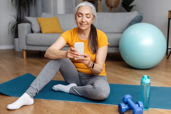 Spor giyimli mutlu atletik kadın evde yoga minderinde oturuyor ve modern cep telefonu kullanıyor, güzel fitness mobil uygulamalarını kontrol ediyor, uzayı kopyalıyor.