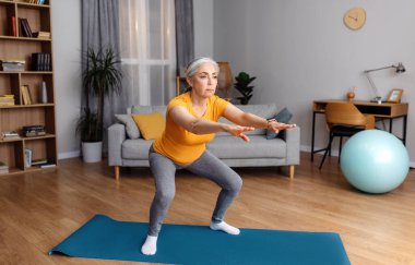 Ev antrenmanı. Son sınıf öğrencisi kadın yoga minderinde çömeliyor, evde çalışıyor, iç mekanda çalışıyor, boş yer var. Formda bir yaşlı kadın ev içi eğitimde bacaklarını çalıştırıyor.