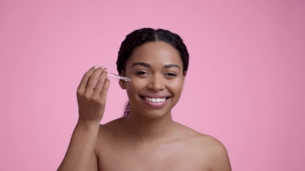 皮肤玻璃化 近距离拍摄年轻快乐的非洲裔美国女人的肖像 用血清涂在脸上 在粉色工作室的背景上笑着拍照 慢动作 — 图库视频影像