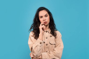 Üzücü, düşünceli, zeki, Avrupalı, gündelik esmer kız öğrenci problemi çözmeyi düşünüyor, mavi arka planda, stüdyoda, boş alanda izole edilmiş. Beyin fırtınası, el işareti, yüz ifadesi, seçim