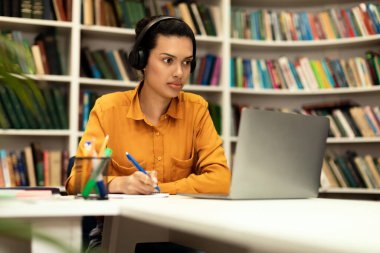 Genç melez bayanın online dersi var, kütüphanede dizüstü bilgisayarın önünde oturuyor, kulaklık takıyor ve notlar alıyor, e-eğitimin tadını çıkarıyor, kopyalama alanının tadını çıkarıyor.