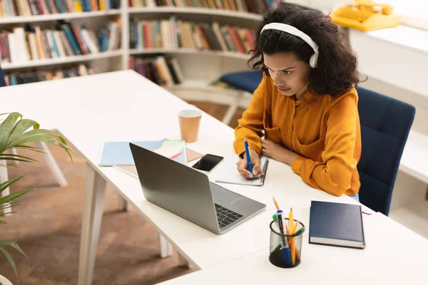ノートパソコンでオンラインレッスンを受けるヘッドフォンの黒人女性に焦点を当て 図書館に座っている間にノートブックで書く 若い女子学生がメモを取り遠隔で勉強し — ストック写真