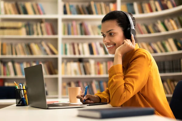 現代のテクノロジーとEラーニング オンラインでウェビナーを見て 図書館に座って ノートパソコンを使用してノートを取る フリースペースで幸せな混合レースの女性 — ストック写真