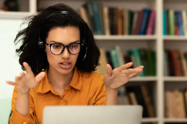 Modern remote education. Female teacher having videochat with students, explaining new materials while talking to laptop webcam, sitting in library interior