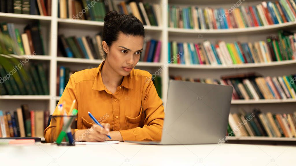 Focused woman using laptop and writing in notebook, studying online in library interior, browsing internet, watching webinar or lecture, panorama with free space