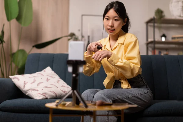 化粧品や化粧品を使って動画を作る韓国のプレッシャーブロガーの女性が自宅でソファに座って携帯電話で撮影します 美容ブログのコンセプト 選択的フォーカス — ストック写真