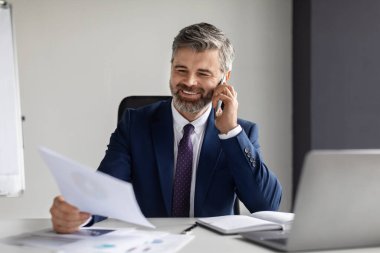 Gülümseyen orta yaşlı işadamı cep telefonuyla konuşuyor ve ofisteki kağıtları kontrol ediyor, yakışıklı, olgun girişimci mali raporları okuyor ve iş ortağına telefon ediyor, mekanı kopyalıyor.