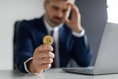 Kripto Hatası. Ofiste dizüstü bilgisayarla otururken Bitcoin tutan kızgın işadamı, depresif erkek girişimci, Ümitsiz Başa Dokunma, Çevrimiçi Ticaret 'te para kaybı