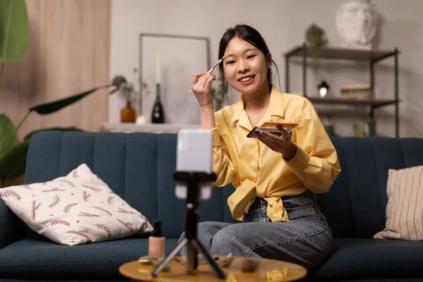 Asian Female Beauty Blogger Making Makeup Filming On Smartphone Sitting On Sofa At Home. Lady Creating Blog Advertising Cosmetics Products. Professional Blogging Concept. Selective Focus