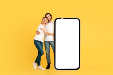 Beyaz tişörtlü neşeli beyaz adam ve kadın sarılıyor, sarı arka planda izole edilmiş, boş ekranlı, dev bir telefonun yanında duruyor. Yeni uygulama, web sitesi ve blog. Harika dijital reklam ve teklif