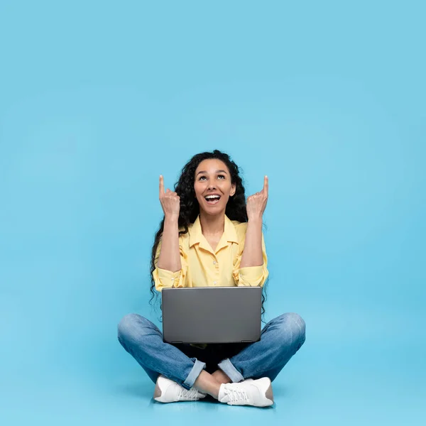 幸せな若い女性がオンラインで作業し クロスレッグに座ってラップトップを使用し 青いスタジオの背景にある空のスペースを指しています 楽しいです千年の女性とともにPc提供場所のための広告 — ストック写真