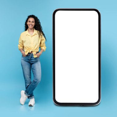 Neşeli genç esmer kadın mavi stüdyo arka planında boş ekranla büyük bir akıllı telefonun yanında poz veriyor. Reklam, mobil uygulama ya da yeni web sitesi için bir model. Boş boşluklu cep telefonu görüntüleme şablonu